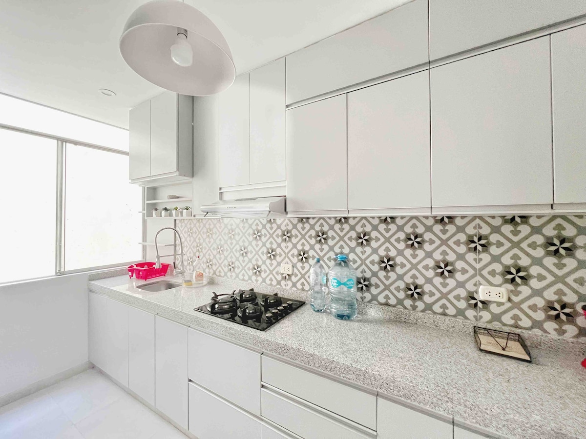 A modern kitchen is equipped with white cabinetry and a patterned backsplash. Natural light comes through large windows, enhancing the workspace. A stove and a water jar are visible on the countertop, complemented by minimalist decor and ample storage.