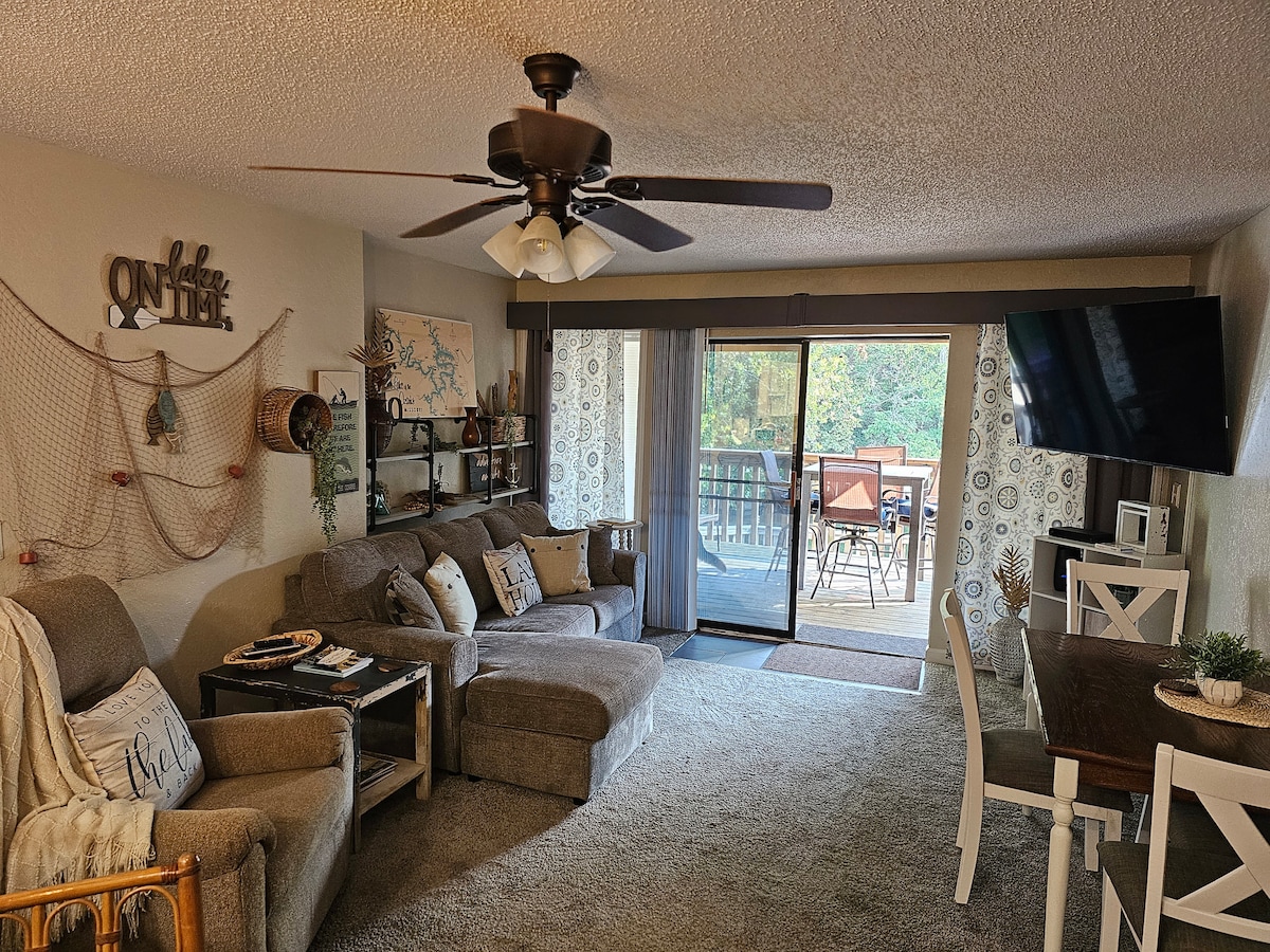The living room features a spacious couch with a chaise lounge and a recliner, accompanied by end tables. A large sliding glass door leads to the outdoor deck, providing light and views. A 58" TV is mounted on the wall, and a dining table with chairs is also visible.