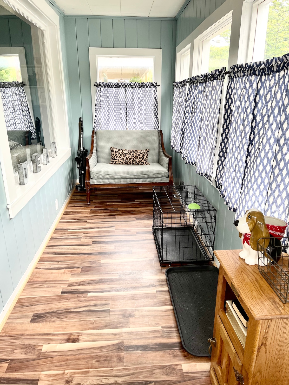 A bright sunroom features large windows adorned with patterned curtains, allowing natural light to fill the space. A comfortable seat with decorative pillows rests against the wall, while two pet crates occupy the floor. A wooden cabinet adds functional storage beside a pet mat.
