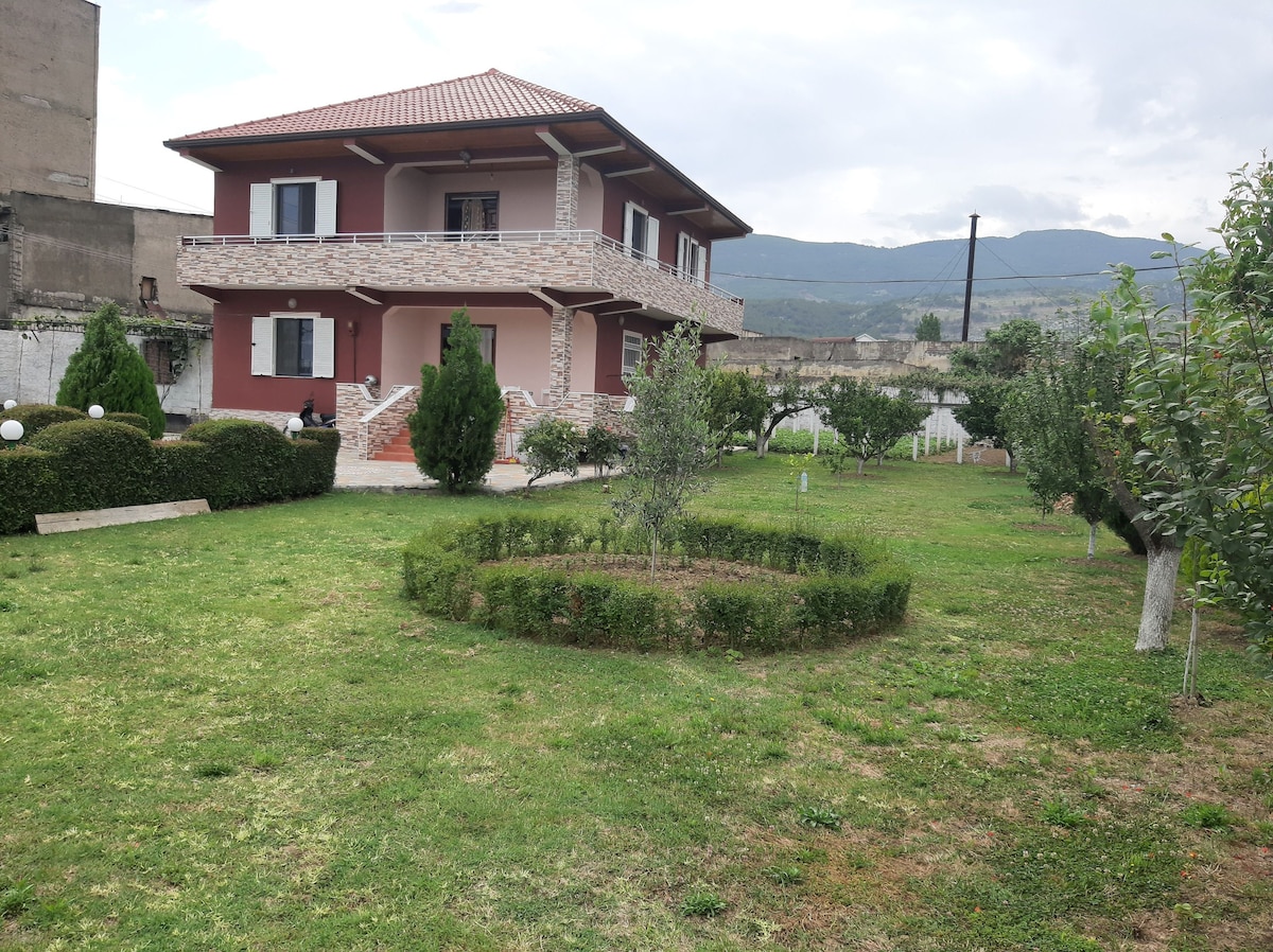 A two-story house is positioned within a spacious garden, featuring a well-maintained lawn and neatly trimmed hedges. Trees are scattered throughout, and the facade displays red and stone accents. A scenic backdrop of rolling hills is visible in the distance under a cloudy sky.