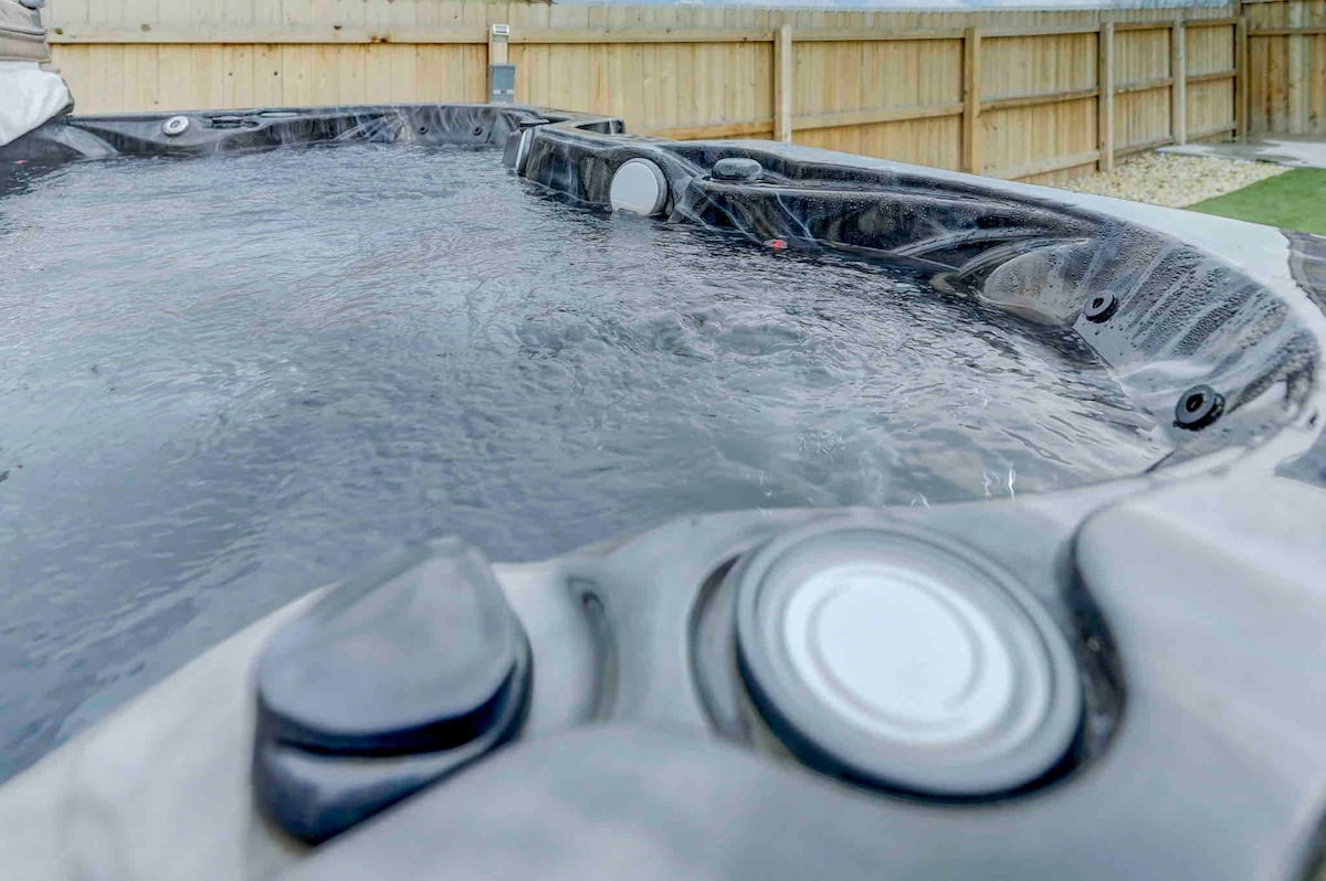 A hot tub is shown with a calm water surface, highlighting the circular jets along the edge. The surrounding fenced area ensures privacy, with grassy ground visible nearby.