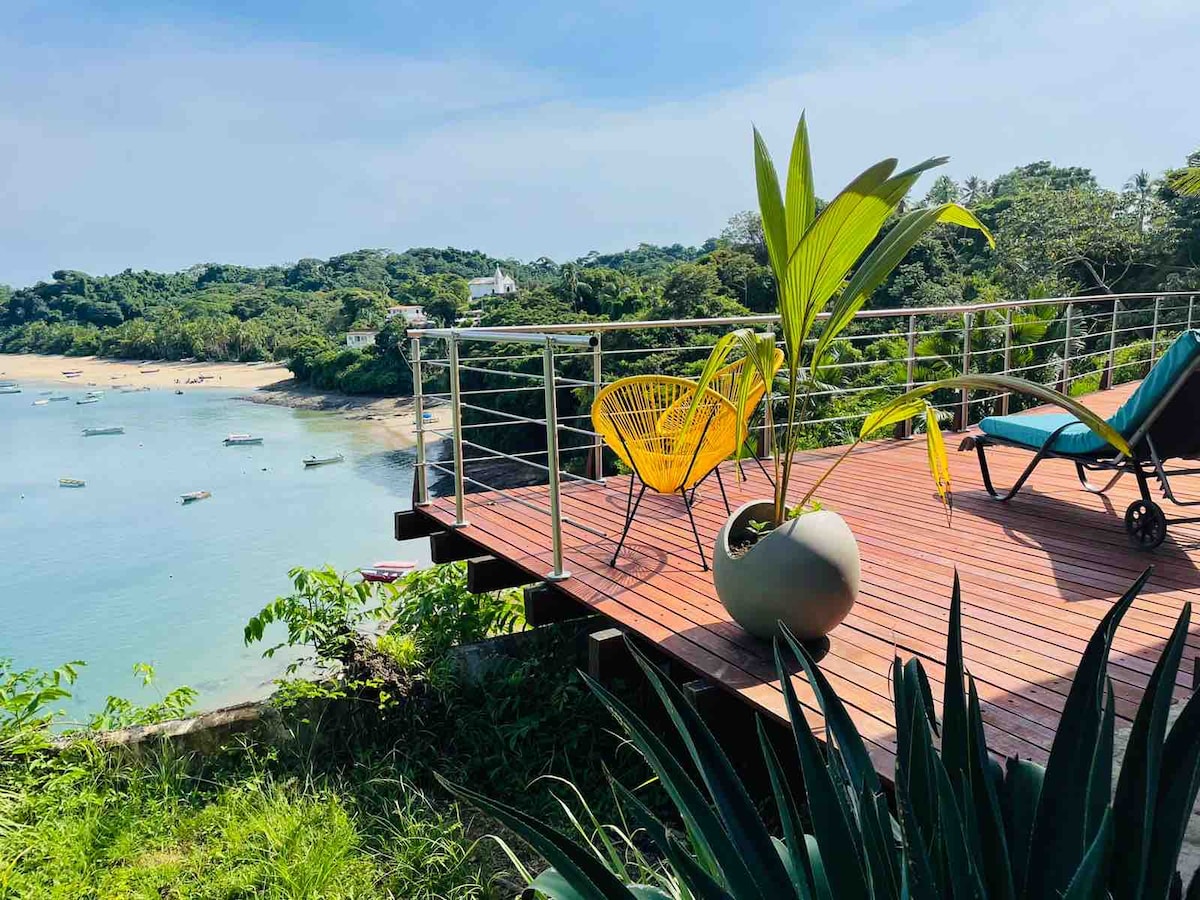 A spacious deck constructed from wooden planks offers a relaxing atmosphere, featuring a vibrant yellow chair and a sun lounger. Lush green vegetation surrounds the area, and a view of the calm bay, dotted with small boats, is visible in the background.