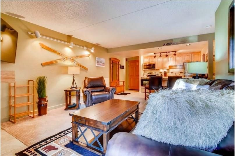 A welcoming living area features a leather sofa adorned with a textured throw, accompanied by a wooden coffee table. A comfortable armchair sits next to a lamp illuminating the space. In the background, a fully equipped kitchen is visible, adding to the room's inviting atmosphere.