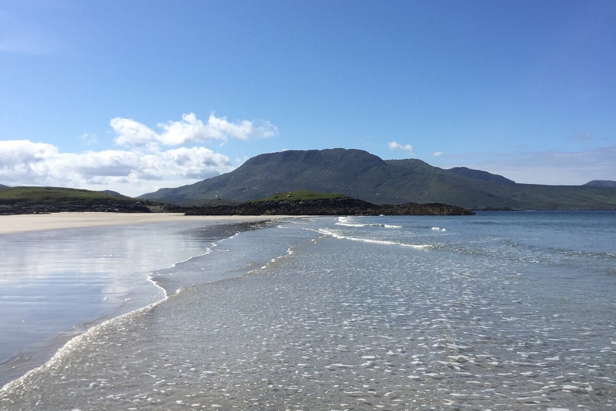 A scenic view of a sandy beach stretches into the distance, bordered by gentle waves lapping at the shore. A mountain stands prominently in the background under a clear blue sky, with scattered clouds adding texture to the horizon.