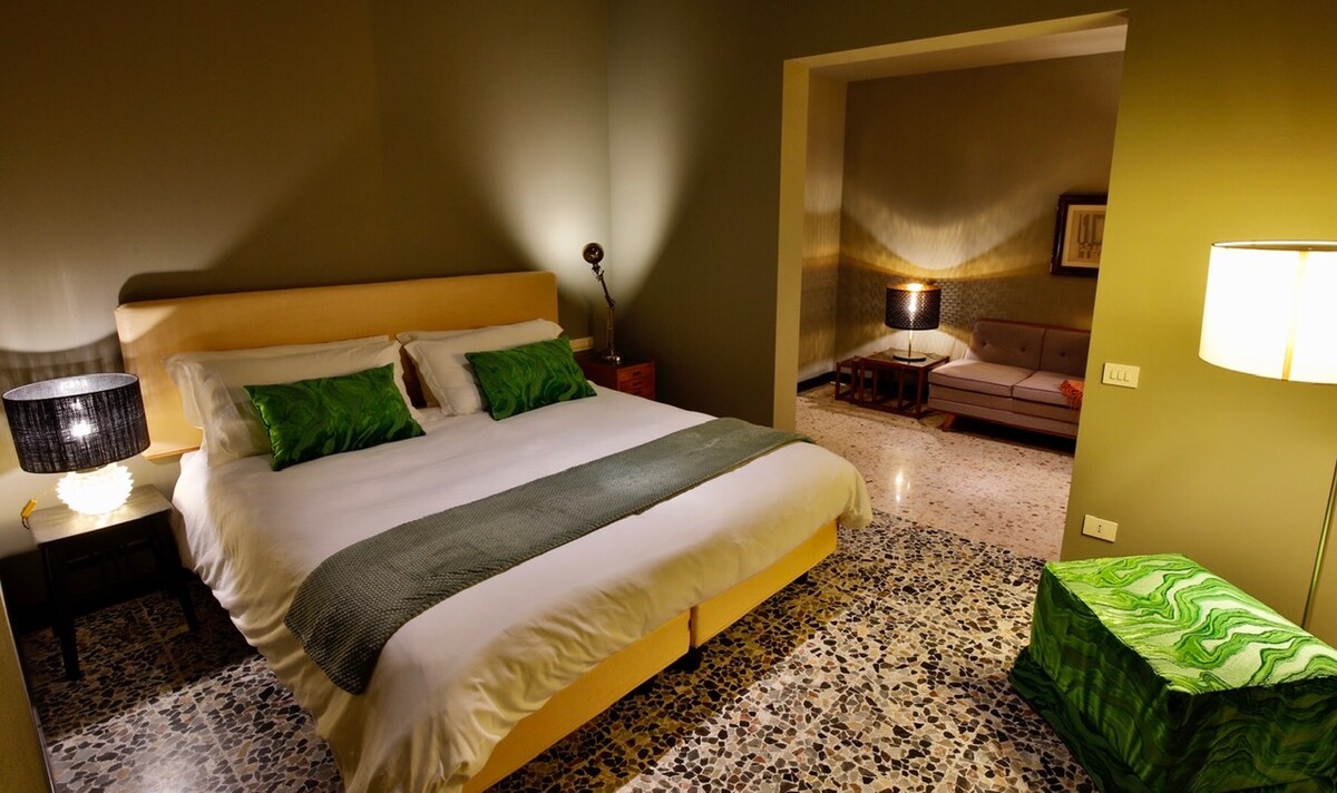A spacious guest room features a large bed with soft linens, accented by vibrant green throw pillows. A bedside lamp provides gentle lighting. A patterned floor adds character, while a separate seating area with a sofa is visible in the background.