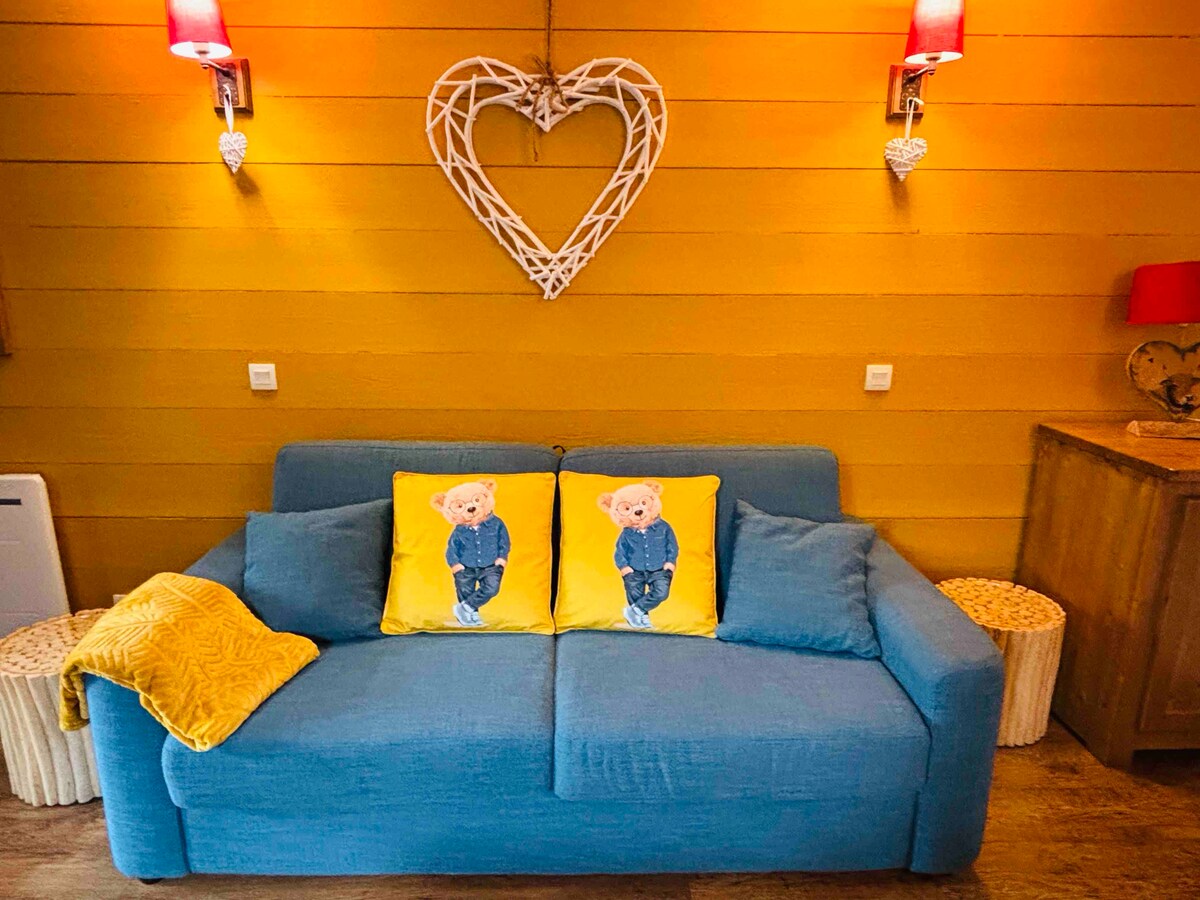 A comfortable blue sofa is adorned with two yellow cushions featuring playful bear designs. The wall behind has a heart-shaped decoration, complemented by two red bedside lamps. Light wood accents and side tables enhance the cozy ambiance of the seating area.