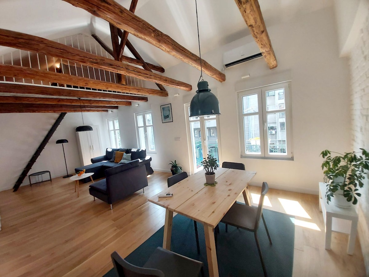 An open living area is highlighted by high ceilings and exposed wooden beams. A wooden dining table is surrounded by chairs, with natural light streaming through multiple windows. A dark sofa and floor lamp add to the inviting atmosphere of the space.
