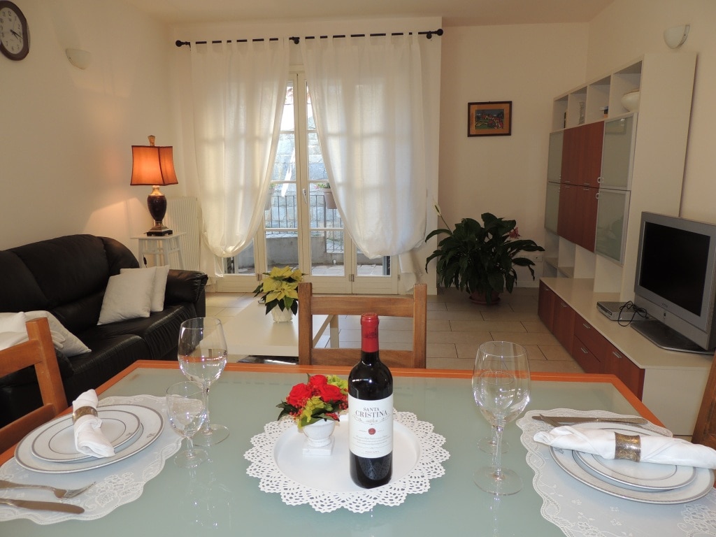 A dining area features a glass table set with plates and wine glasses, complemented by a floral centerpiece. In the background, a comfortable black sofa and a modern TV unit are visible, with sheer curtains framing an outdoor view.
