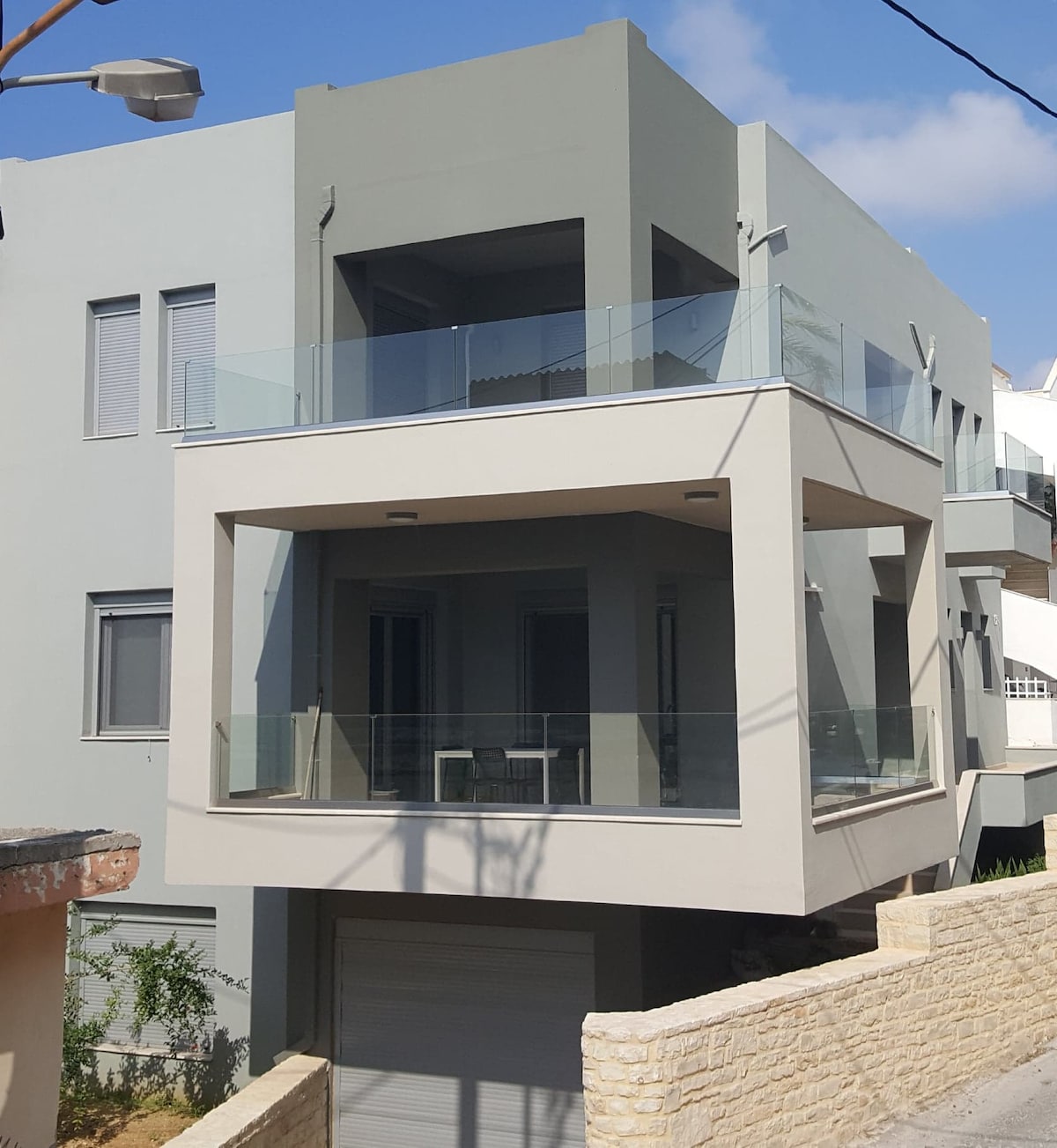 A modern two-storey villa features a prominent balcony with glass railings, providing a view of the surroundings. The light grey exterior is complemented by large windows. The garage entrance can be seen at the base of the villa, surrounded by a stone wall.