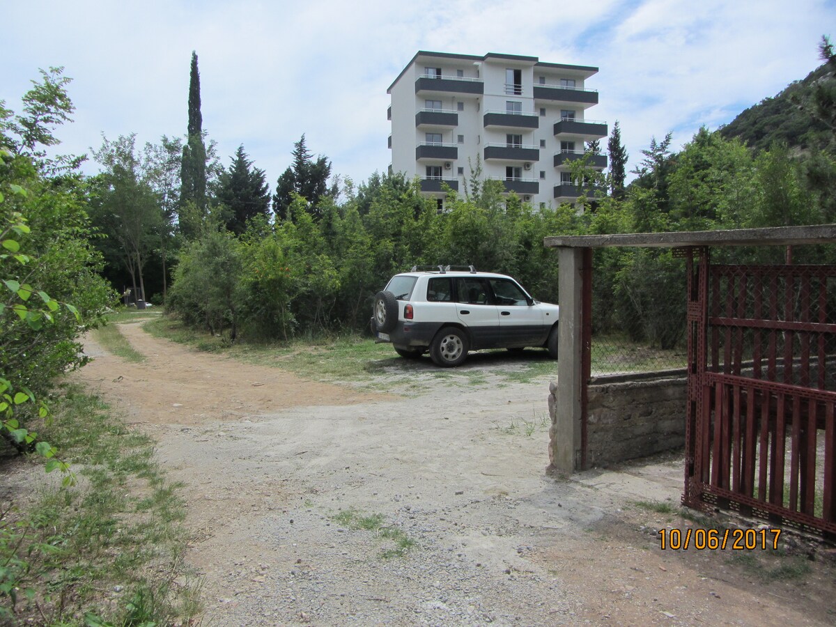 A pathway leads to a gravel parking area surrounded by lush greenery. An SUV is parked nearby, with a multi-story building visible in the background. The scene captures a sense of space and tranquility, with ample room for vehicles.