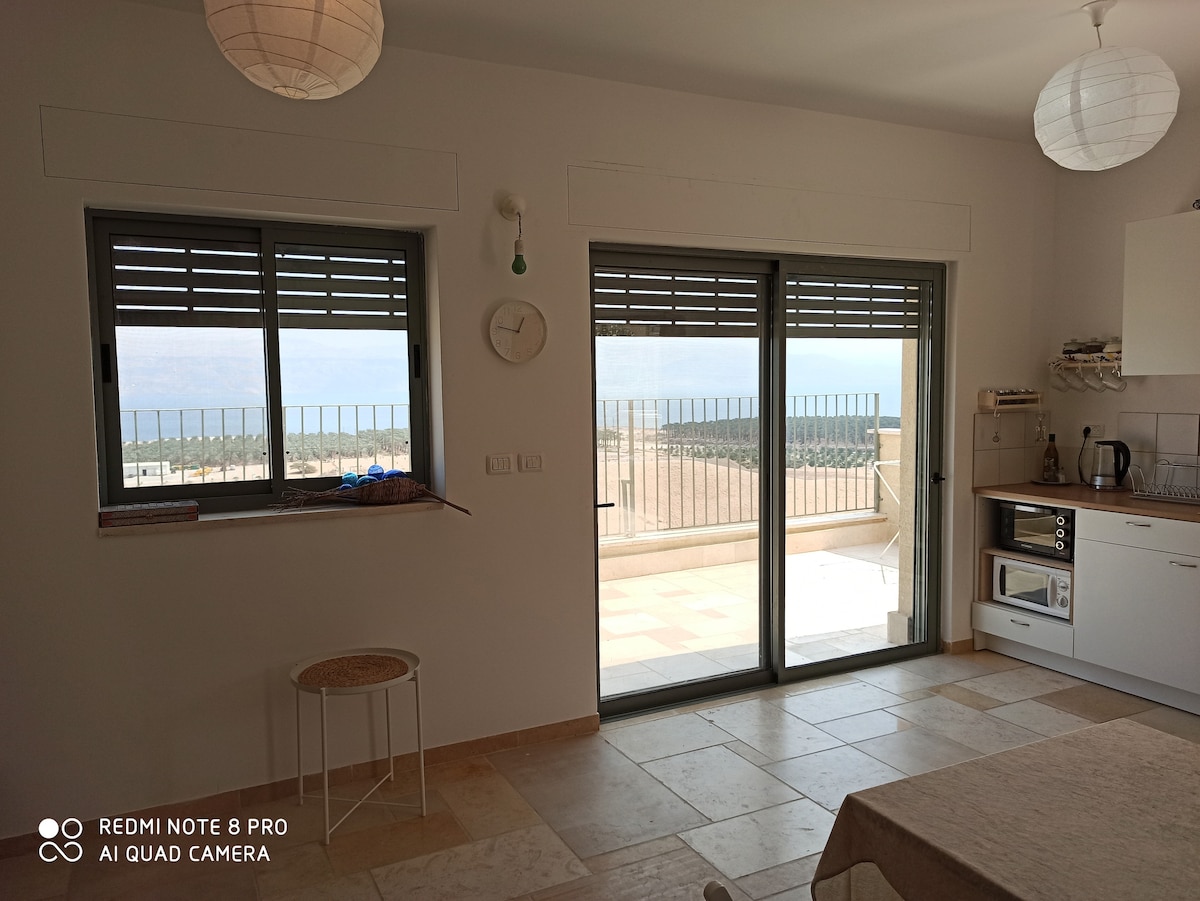 A bright kitchen area features large sliding glass doors that provide access to a balcony with scenic views. Natural light fills the space, highlighting the simple decor, including a wall clock and paper lanterns. A light-toned table stands ready for dining.