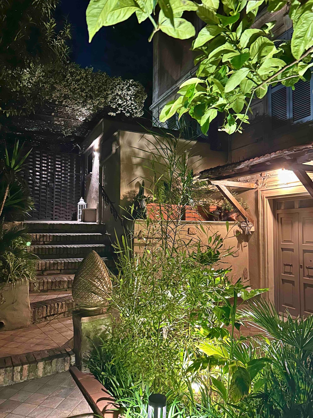 The entrance features a stone staircase illuminated by soft lighting, surrounded by lush greenery and plants. A decorative element is visible near the base of the stairs, while a wooden lattice structure partially obscures the view of the upper level.