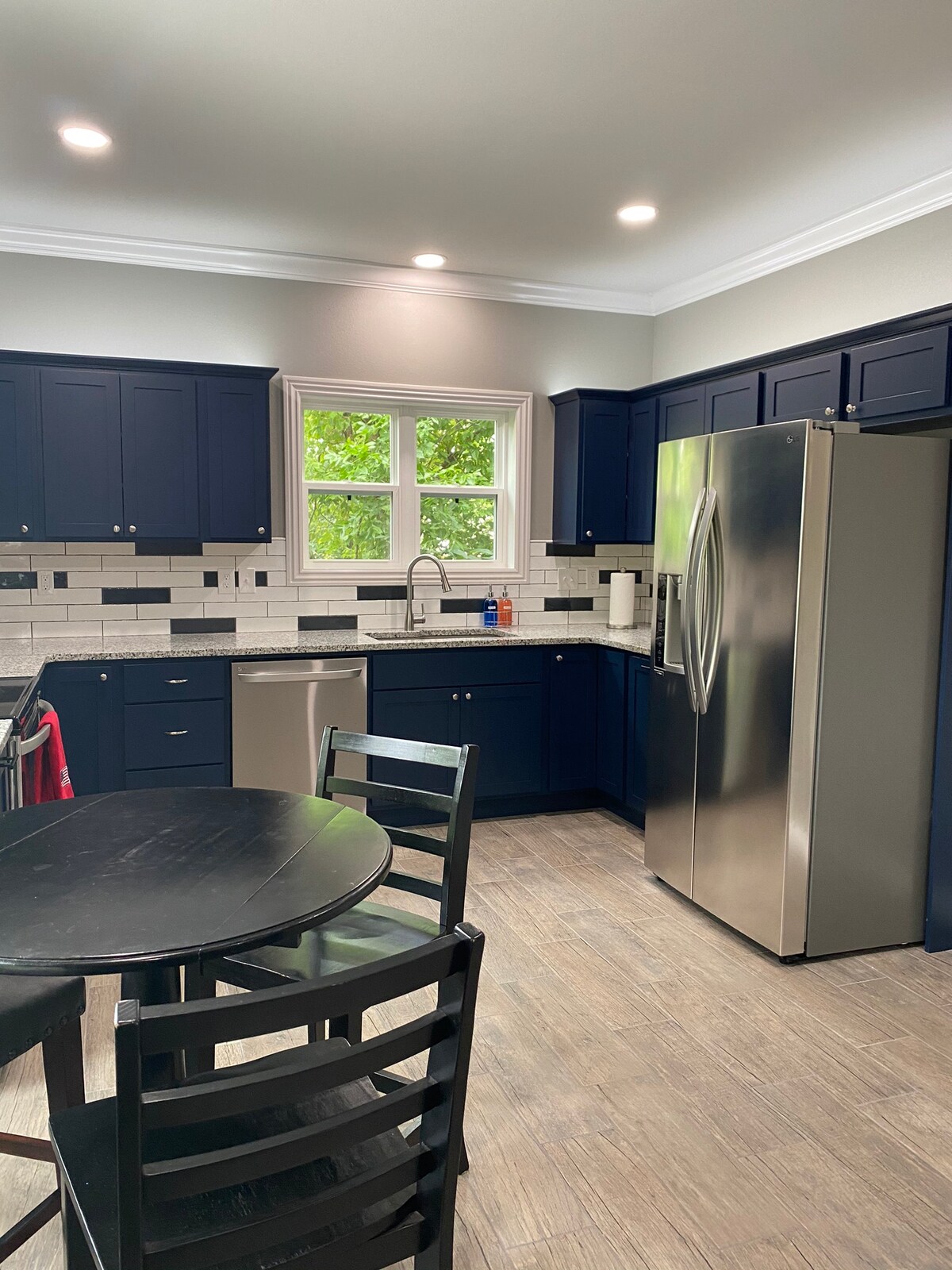 The kitchen features navy blue cabinetry with white subway tile backsplash. A stainless steel refrigerator and dishwasher complement the granite countertops. A round black dining table with four chairs is positioned near a window, allowing natural light to brighten the space.