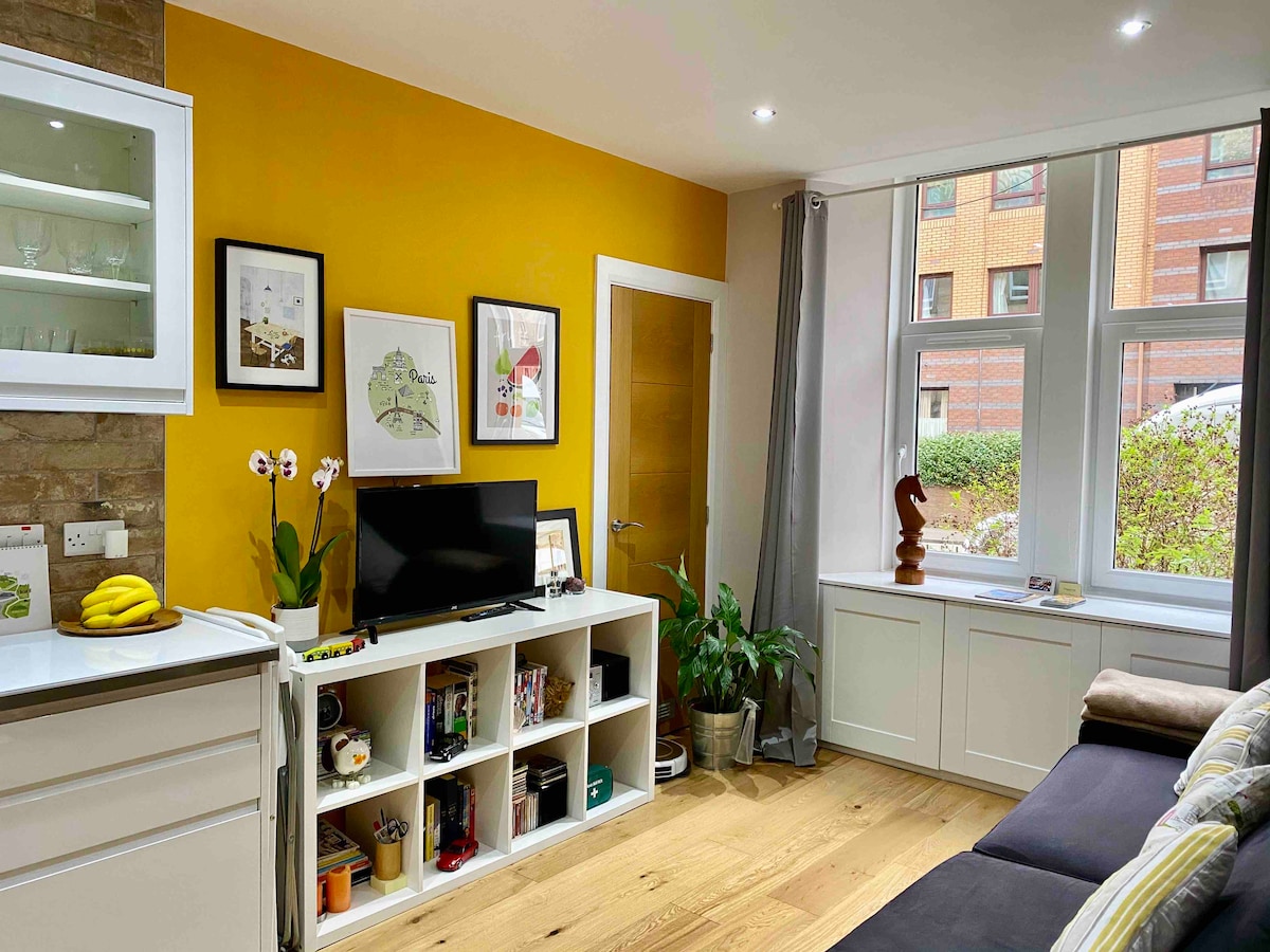 A living area features a warm yellow accent wall and a large window allowing natural light. A television is positioned on a white shelving unit alongside decorative plants. Various framed artworks are displayed on the wall, complementing the modern decor.