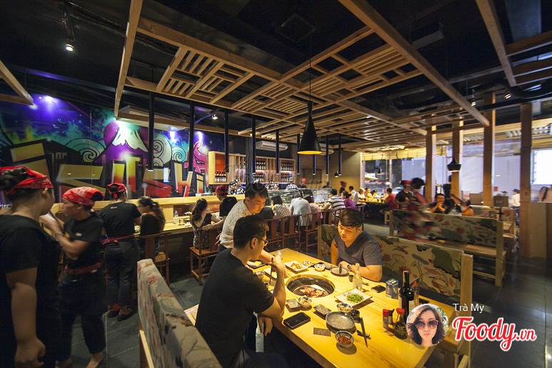 The restaurant interior features an open layout with wooden beams and vibrant wall art. Round tables are filled with diners enjoying their meals, while attentive staff move between tables. A well-lit atmosphere is complemented by decorative lighting fixtures, enhancing the social dining experience.