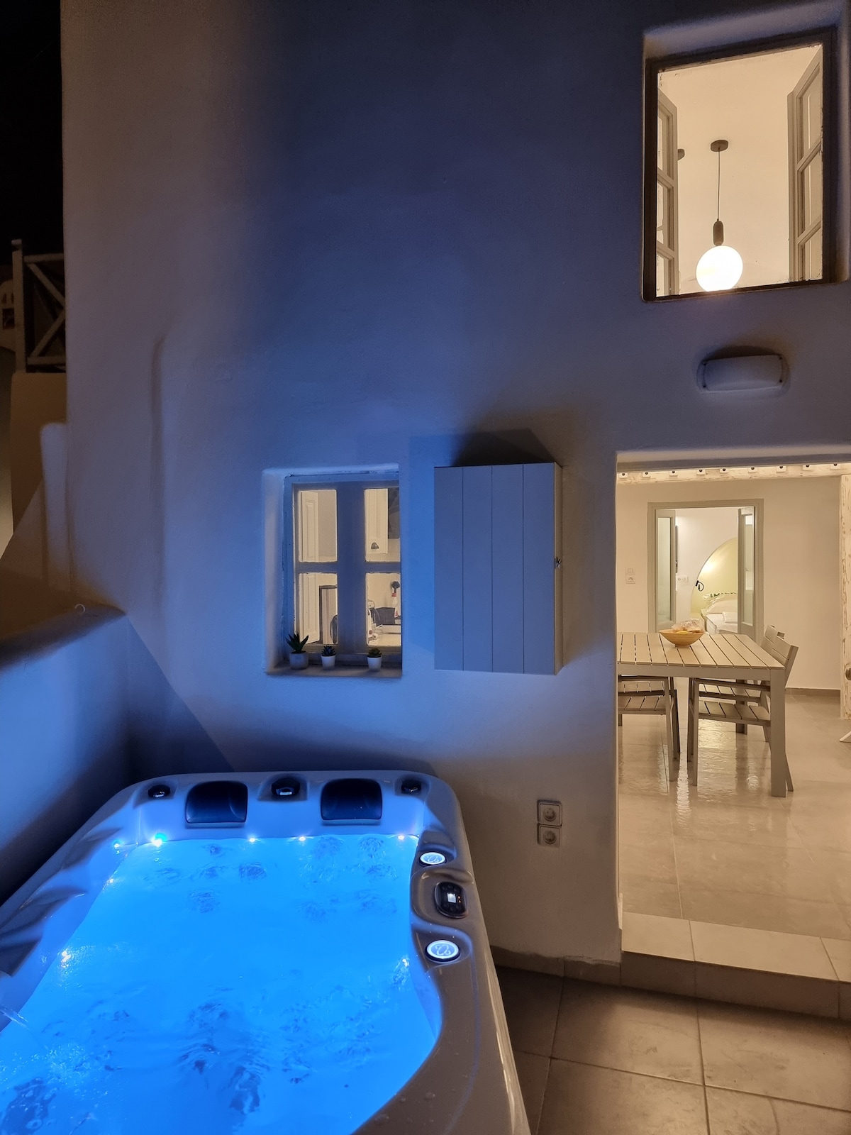 An outdoor patio area features a hot tub, illuminated in soft blue light, creating a relaxing ambiance. To the side, windows reveal a glimpse of the indoor dining space, while a neutral color palette enhances the tranquil setting.