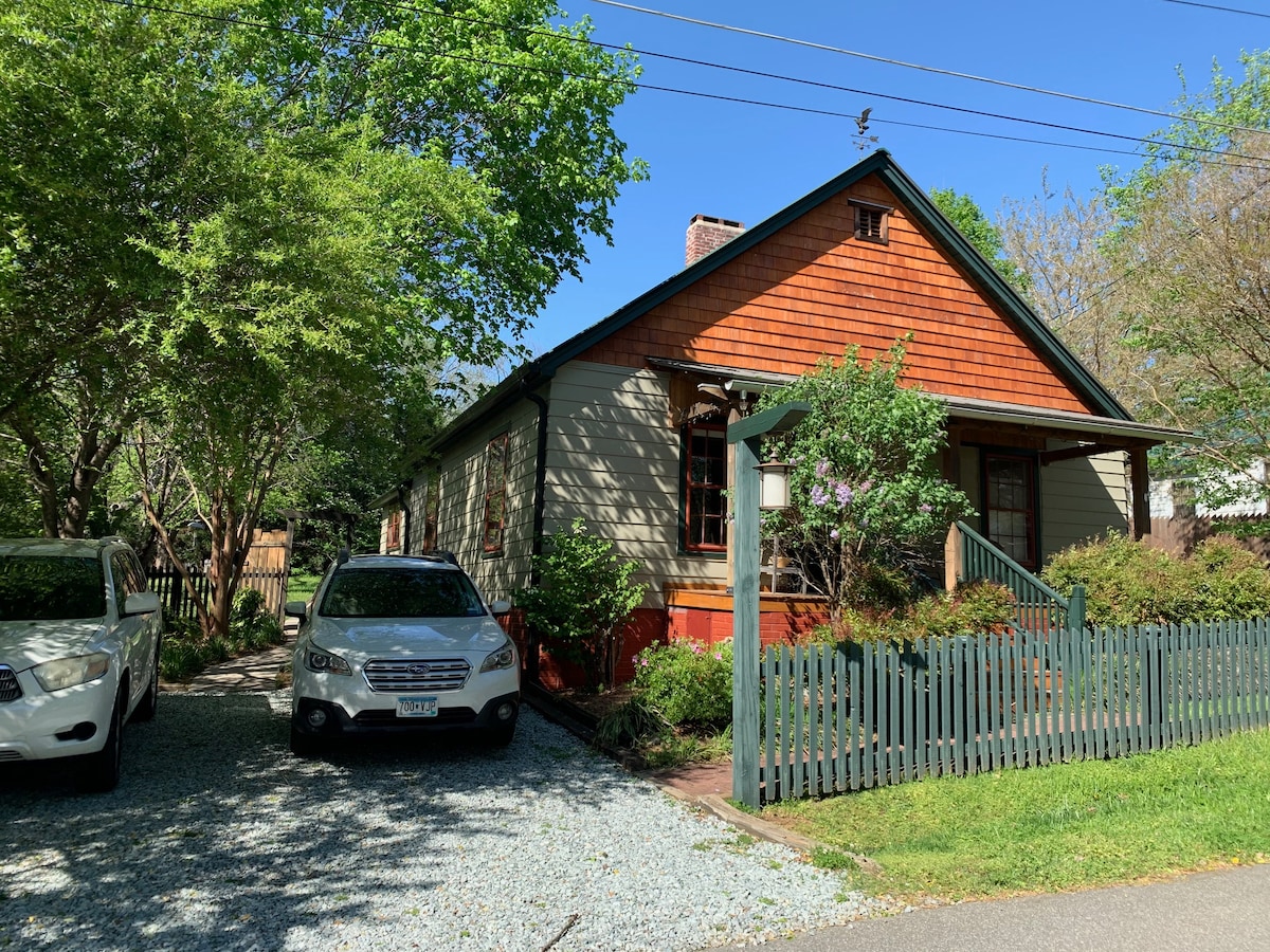 A charming historic home is framed by lush greenery, with a gravel driveway leading to parking. The exterior features a combination of wooden siding and a shingled roof. A picket fence encloses the front yard, enhancing the inviting appearance of the property.