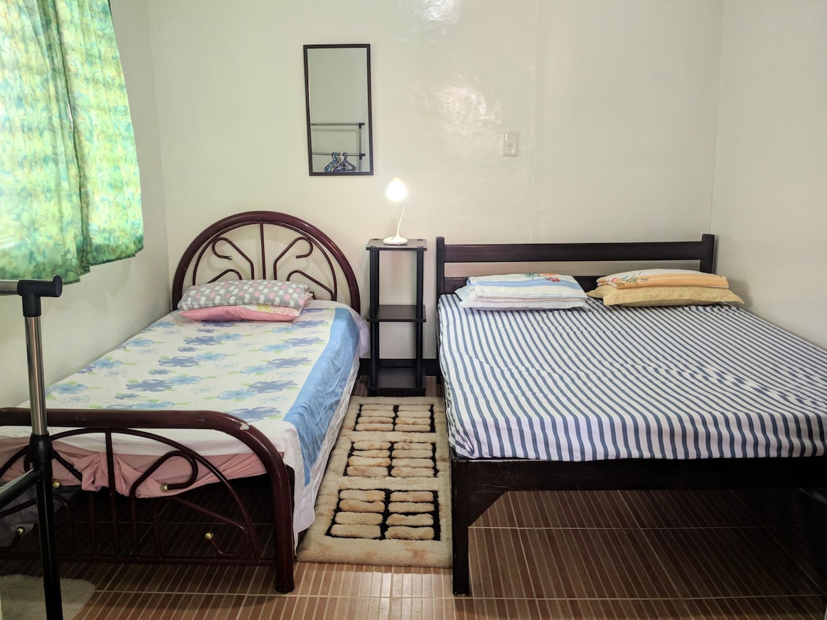 A private room contains two beds with simple bedding in soft colors. A small lamp on a nightstand provides light. A mirror is mounted on the wall opposite the beds. A patterned rug adds warmth to the hardwood floor, and a window covered with a green curtain allows natural light.