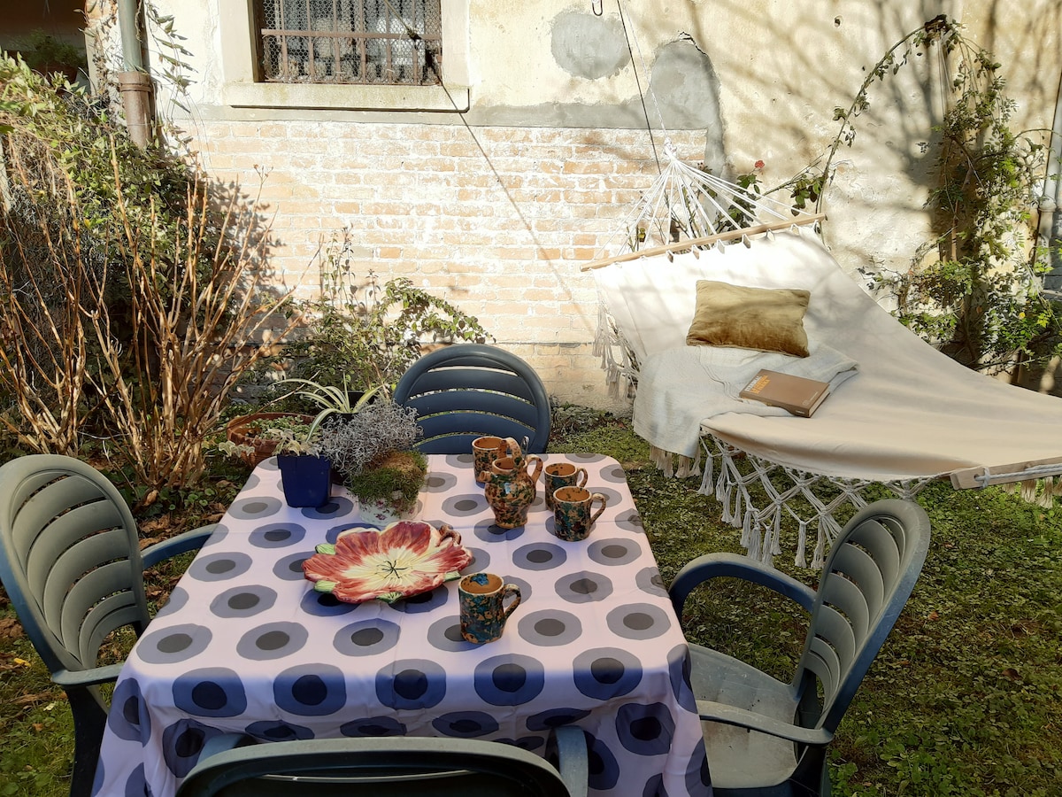 An outdoor garden space features a table set with a floral centerpiece and colorful pottery cups. A hammock is positioned nearby, surrounded by greenery and soft sunlight filtering through trees. The rustic brick wall adds character to the tranquil setting.