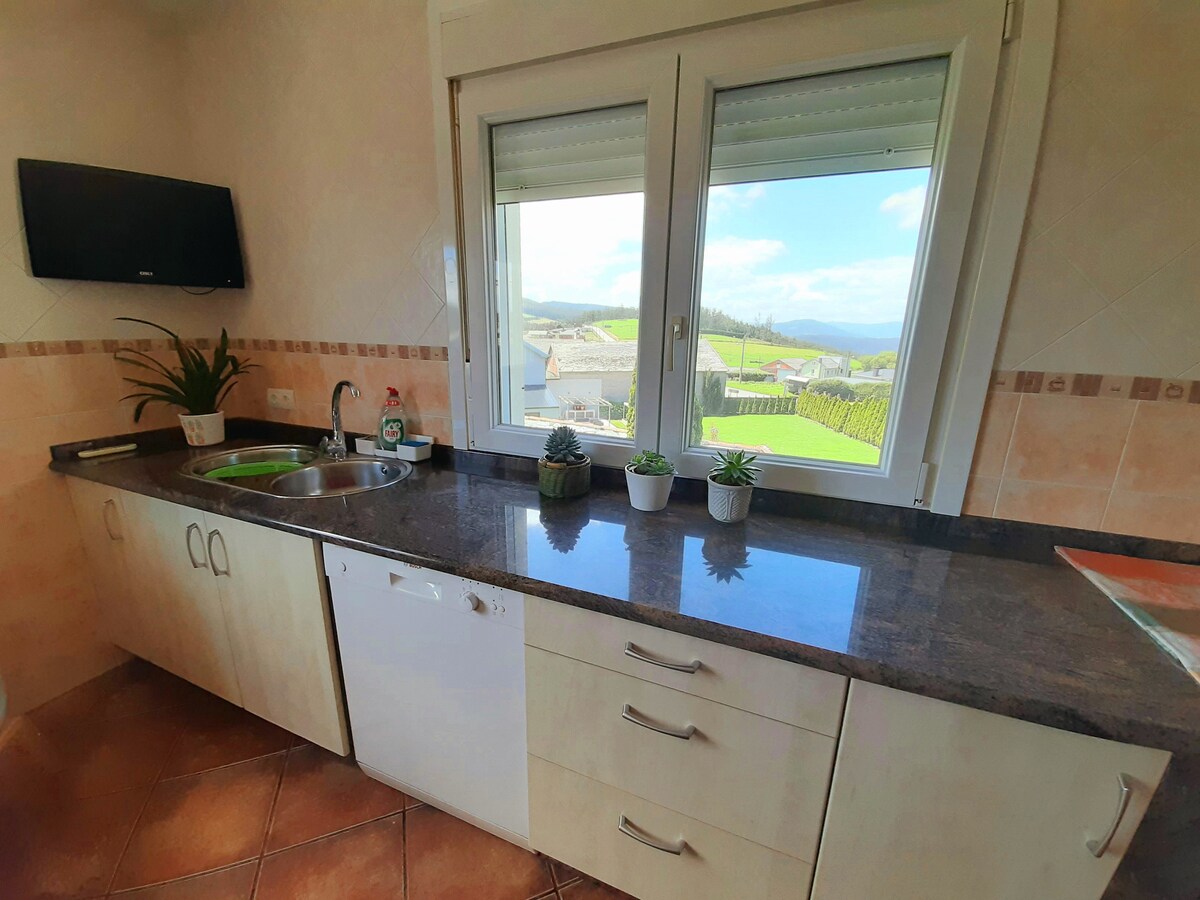A modern kitchen displays a sleek countertop paired with a sink and dishwasher. Large windows provide a view of the surrounding countryside, with greenery visible outside. A television is mounted on the wall, and small potted plants add a touch of greenery.