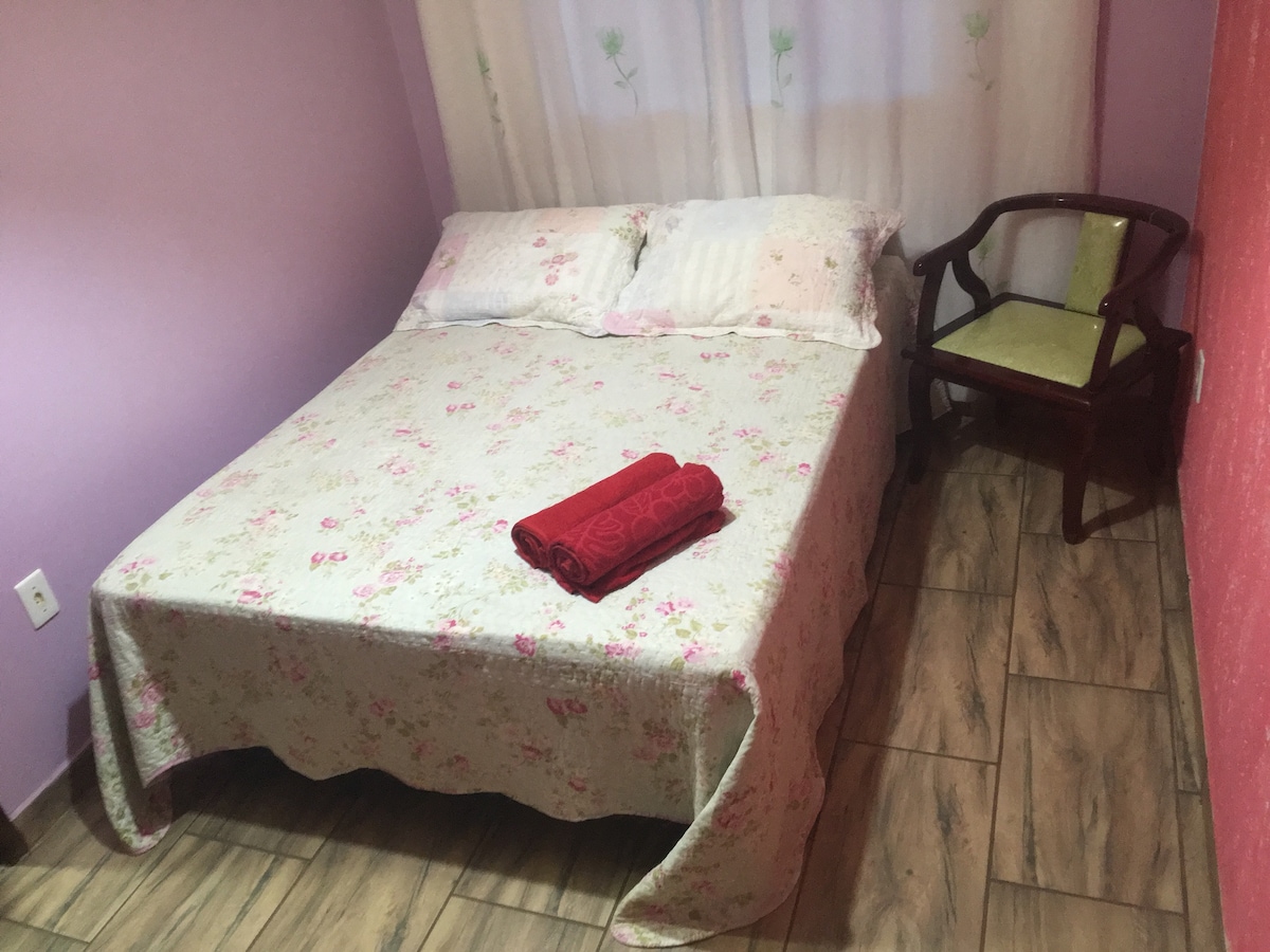 A compact bedroom features a floral-patterned bedspread draped over a spacious bed, complemented by two pillows. A wooden chair with a light green cushion is positioned beside the bed. Natural light enters through a window adorned with sheer curtains.