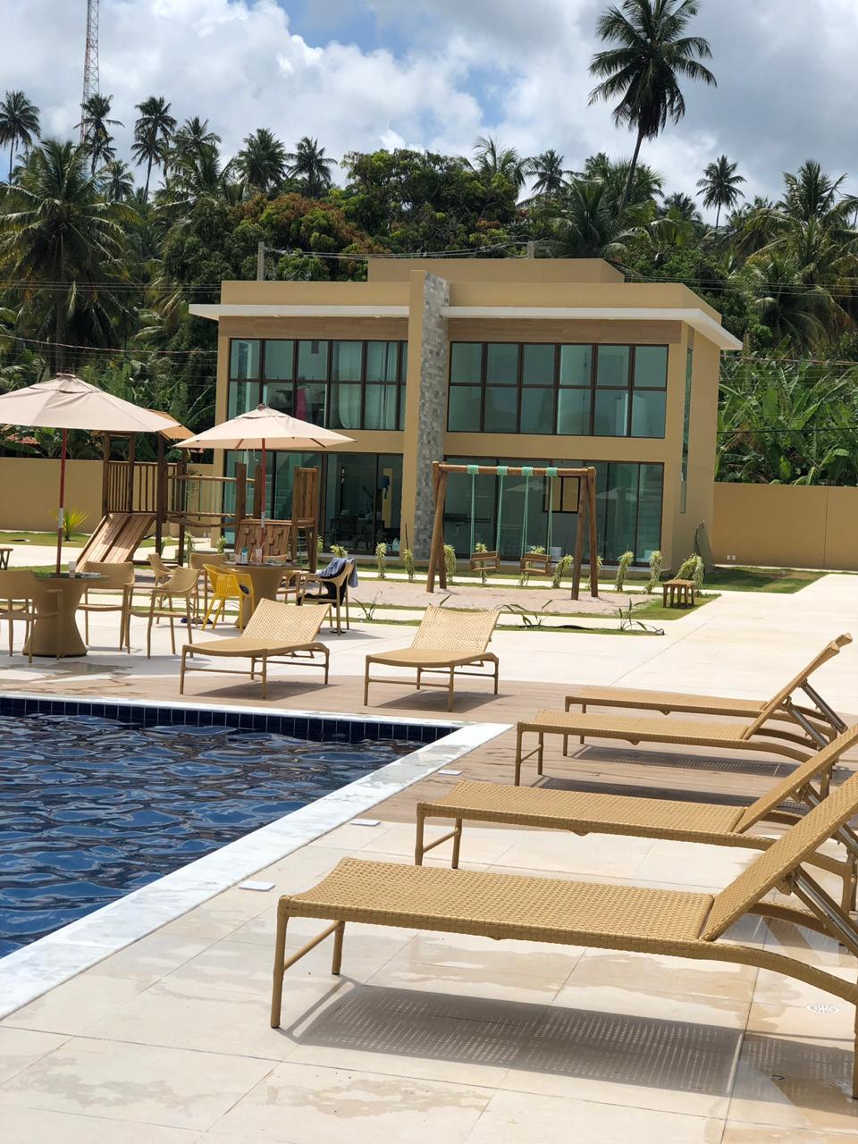 A modern two-story house is visible in the background, featuring large glass windows. In the foreground, a pool is surrounded by sun loungers and umbrellas. Lush greenery and outdoor play equipment are present, creating a relaxing environment.