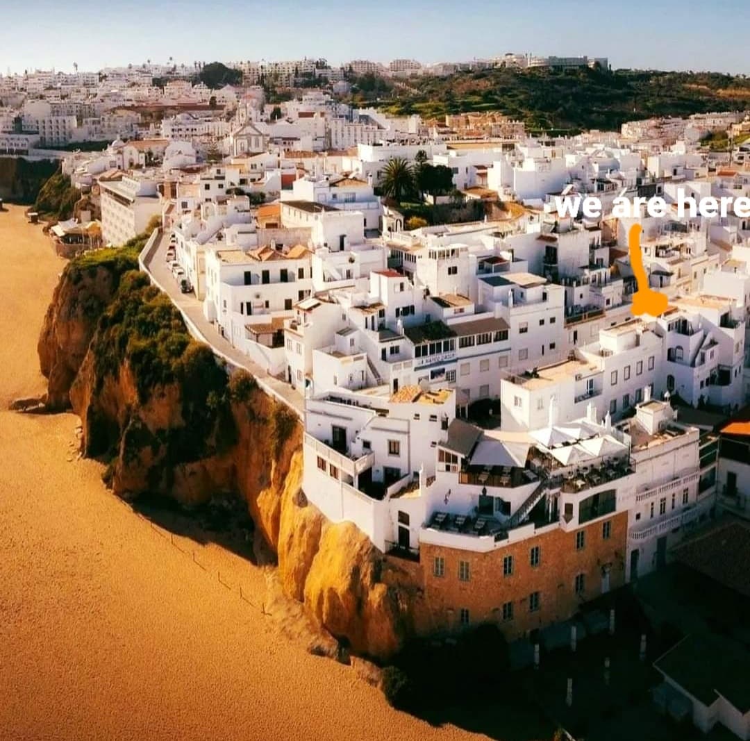 Aerial view of Albufeira's historic center reveals whitewashed buildings nestled along a rocky coastline. The sandy beach curves below, with cliffs providing natural boundaries. The scene illustrates a vibrant area surrounded by greenery, showcasing the charm of traditional Portuguese architecture.