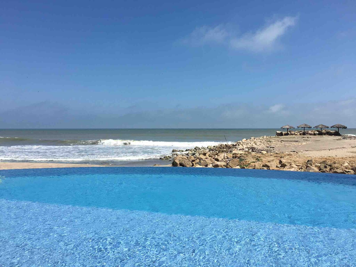 An infinity pool glistens in the foreground, seamlessly merging with the ocean view. Gentle waves lap against the sandy shore, with beach umbrellas visible in the distance under a clear blue sky.