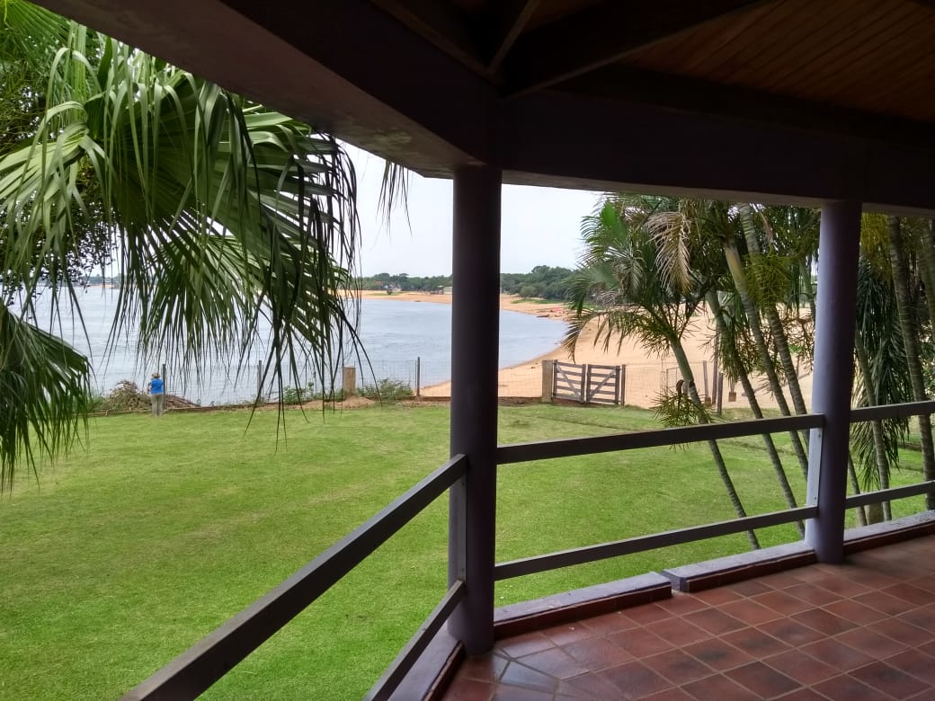 A vantage point from a covered patio showcases a lush green lawn leading to the shoreline of the Paraná River. Tall palm trees frame the scene, and sandy beach areas are visible in the background, providing a serene view of the water.