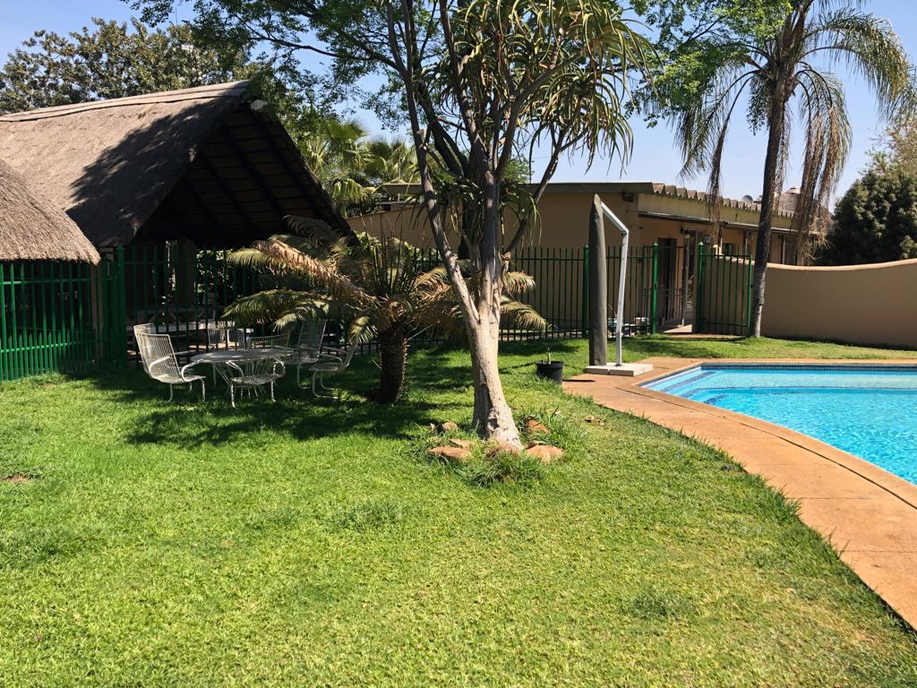 A lush green lawn surrounds a swimming pool, with a shaded seating area featuring a thatched roof. Several chairs and a table are positioned under the shade. Nearby, palm trees and shrubs complement the tranquil outdoor setting.