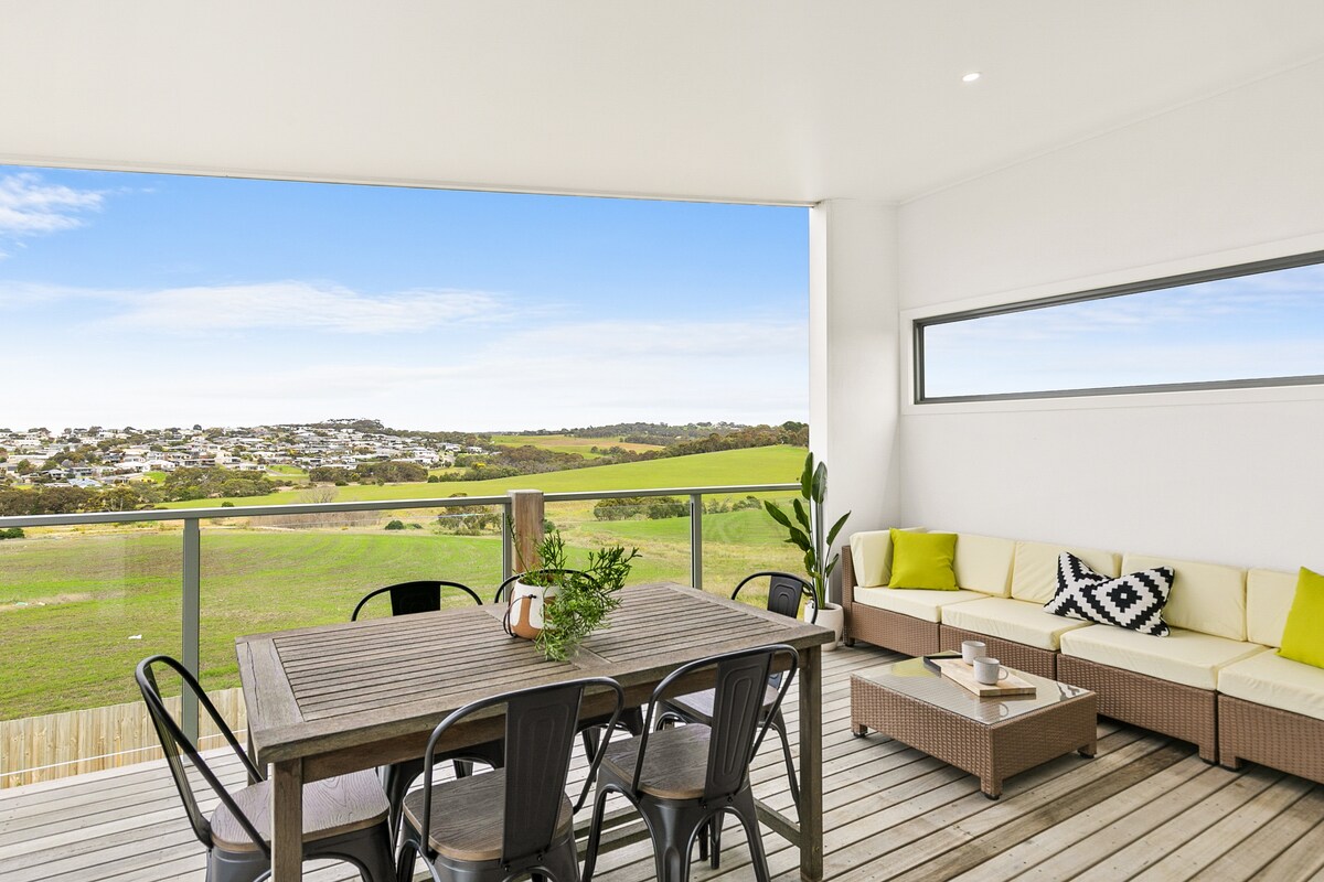 An outdoor deck space features a wooden dining table surrounded by metal chairs, providing seating for several guests. A comfortable lounge area with light-colored cushions is included. The landscape showcases rolling fields and a distant neighborhood, with a clear blue sky overhead.