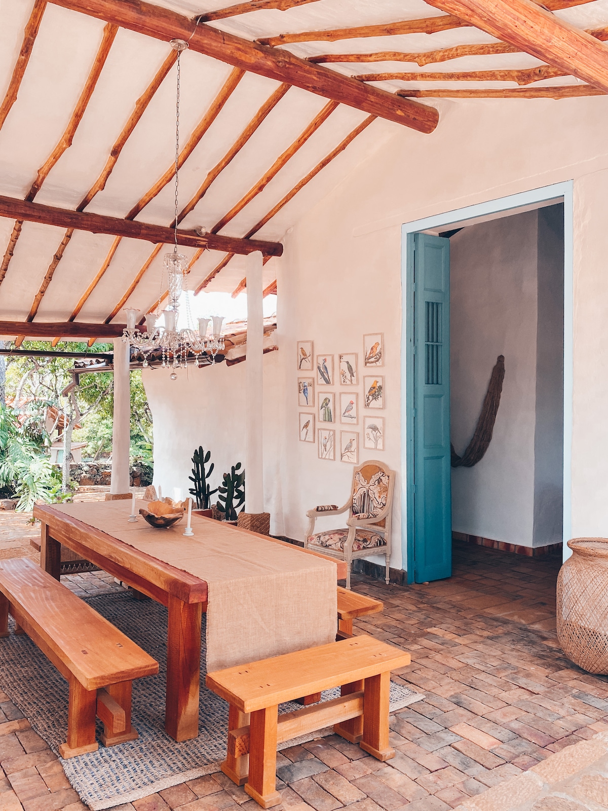 A communal dining area is highlighted by a long wooden table surrounded by benches. Soft light from a chandelier illuminates the space, while woven decor and framed artwork adorn the walls. A doorway leads to another room, enhancing the open atmosphere.