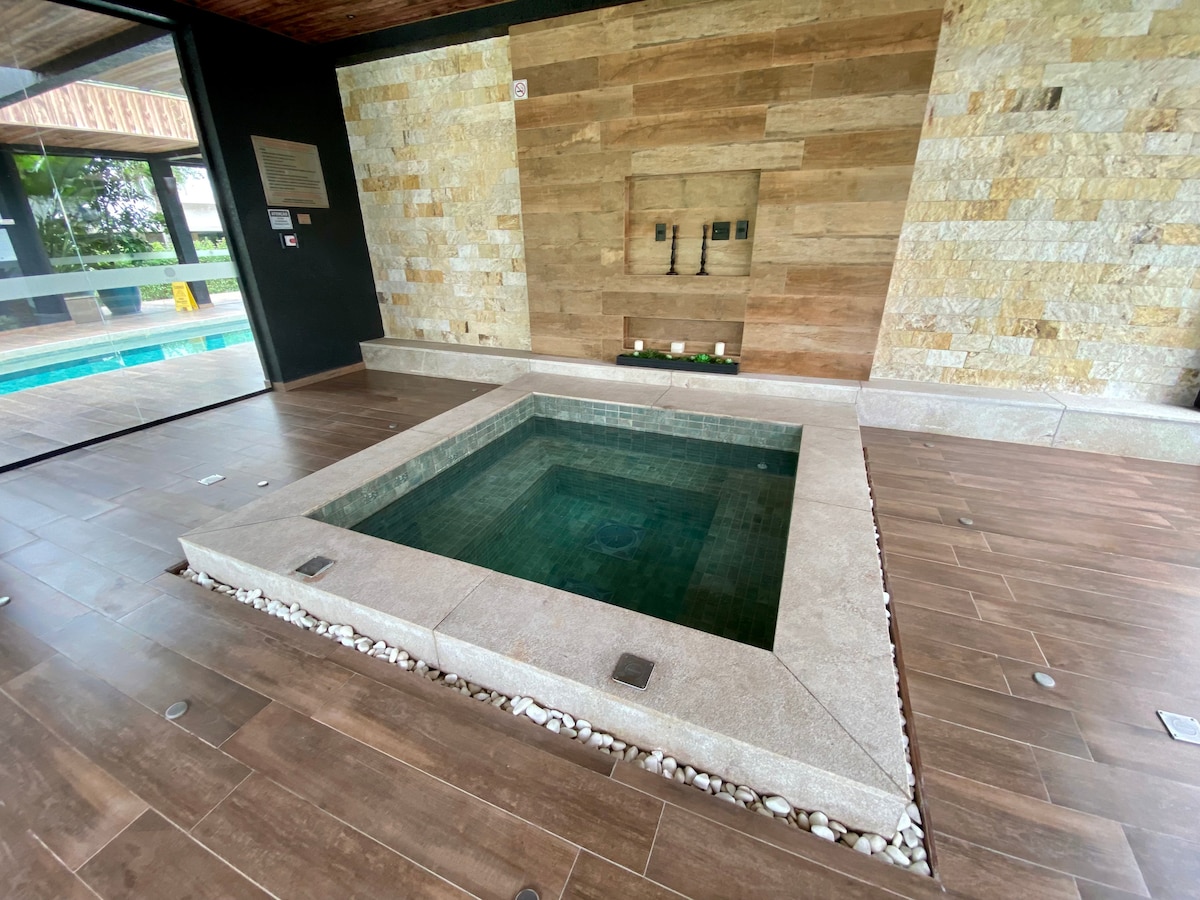 An inviting jacuzzi is positioned in a sleek indoor area. Natural stone walls offer a calming backdrop, while the surrounding floor features warm wooden tiles. Soft lighting highlights the tranquil atmosphere, and a glimpse of the outdoor pool area is visible through large glass doors.