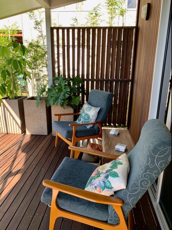 A private balcony is adorned with two comfortable chairs, each featuring decorative pillows. A small wooden table sits between them, surrounded by lush plants and natural wooden elements. Sunlight filters through the space, creating a serene and relaxing outdoor area.