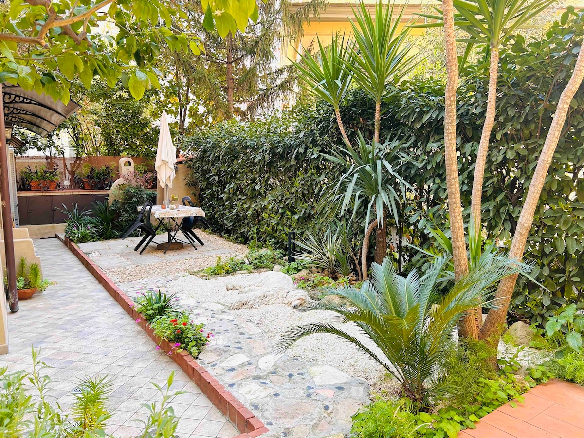A well-maintained garden features a mix of lush greenery and decorative stones. A table with chairs is set under an umbrella, creating a cozy outdoor dining area. Surrounding plants and trees add to the tranquil atmosphere.
