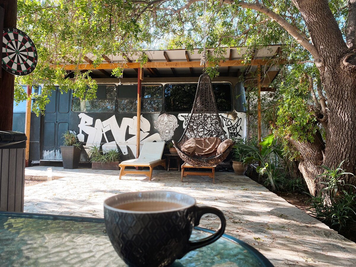A cozy outdoor area features a hanging chair and lounge chairs surrounded by greenery. In the foreground, a cup of coffee is placed on a table. The backdrop includes a mural on the bus and a partially shaded stone patio.