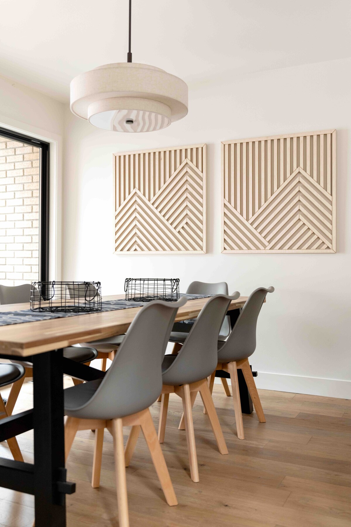A dining area features a long wooden table surrounded by six modern, gray chairs. Above, a pendant light provides illumination. On the walls, two pieces of geometric wall art create a cohesive design, enhancing the minimalist aesthetic.