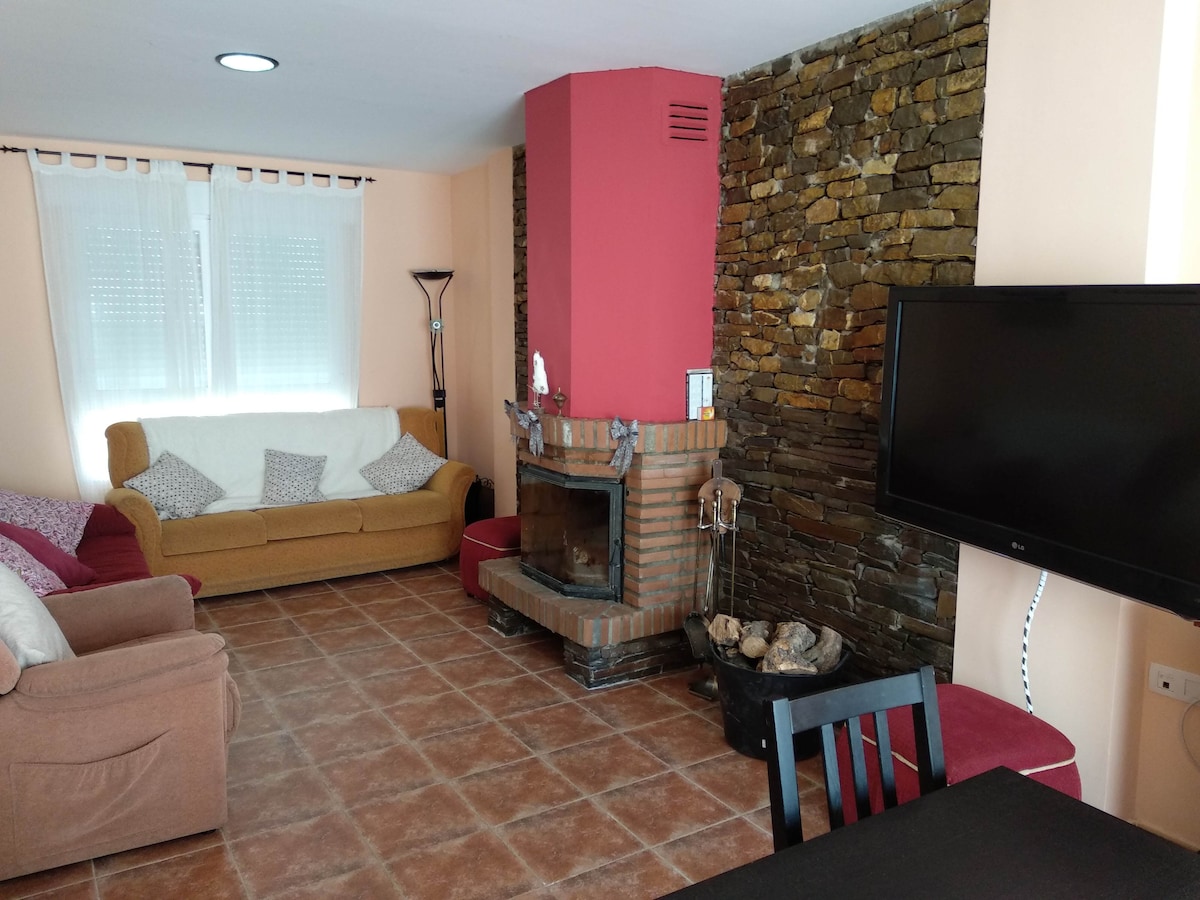 A living room features a stone accent wall, complemented by a red fireplace. A sofa and a chair provide comfortable seating. Natural light enters through a window covered with sheer curtains. A large flat-screen TV is mounted on the wall, enhancing the room's functionality.