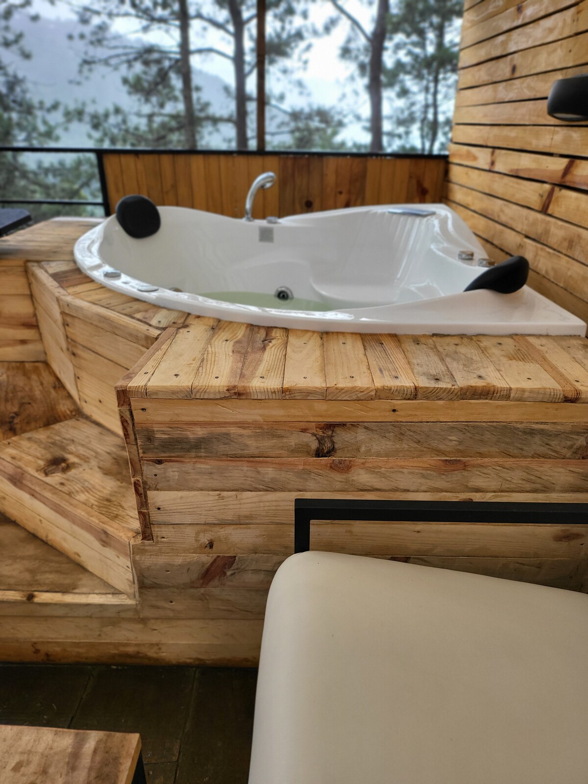 An outdoor jacuzzi surrounded by wooden decking offers a relaxing space. The structure features steps leading up to the jacuzzi, enhancing accessibility, with soft seating nearby for comfort. A view of the natural surroundings is visible through the nearby railing.