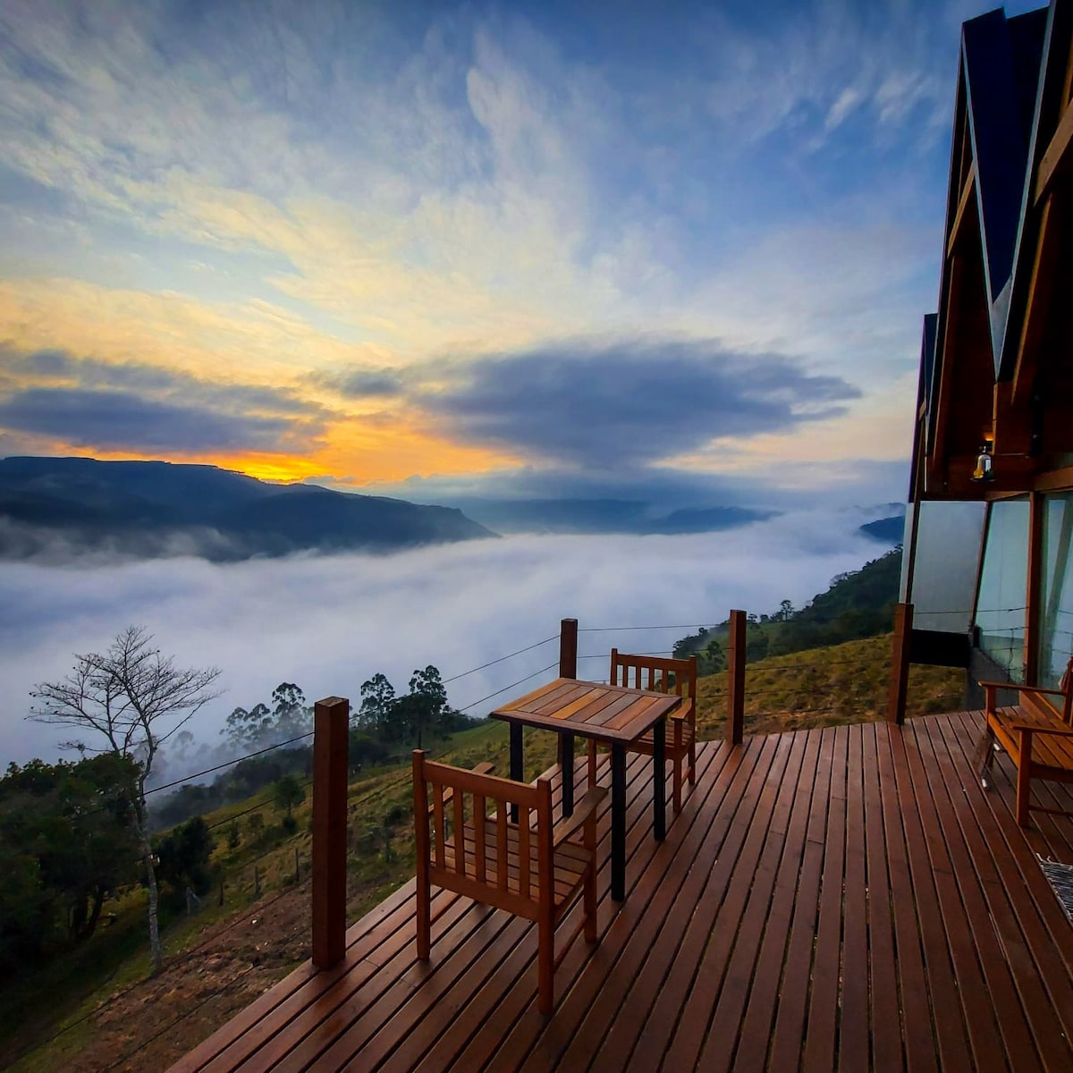 A wooden deck presents a table and chairs set against a backdrop of rolling mountains shrouded in mist. The sky showcases soft pastel hues of orange and blue as the sun rises, creating a serene and inviting outdoor space.