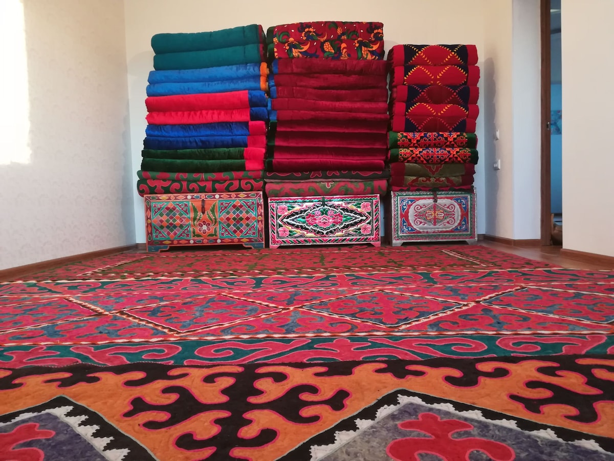 Vibrant patterned rugs cover the floor, showcasing intricate designs in various colors. A stack of neatly arranged blankets in bright hues is displayed against the wall, adding warmth to the space. Subtle textures and detailed patterns are noticeable throughout the room.