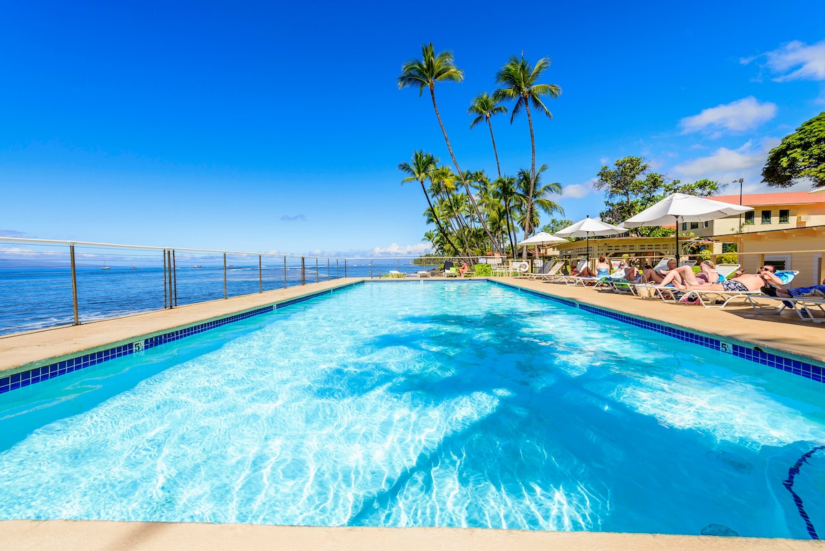 A sparkling oceanfront pool glistens under a clear blue sky, framed by palm trees. Lounge chairs line the poolside, inviting relaxation while enjoying views of the ocean. The tranquil waters reflect the sunlight, creating a serene atmosphere for guests to unwind.