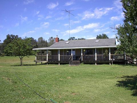 A log home sits on a grassy expanse, featuring a wide front porch with wooden railings. Several trees provide shade in the surrounding landscape. The roof is topped with an antenna, and the clear blue sky enhances the serene country setting.