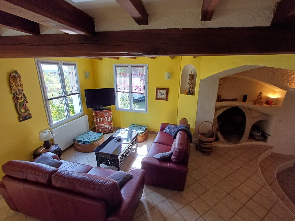 A living area features comfortable seating in rich maroon leather, arranged around a decorative coffee table. Walls are painted warm yellow, and a flat-screen TV is positioned under bright windows with floral curtains. A cozy fireplace adds warmth to the space.