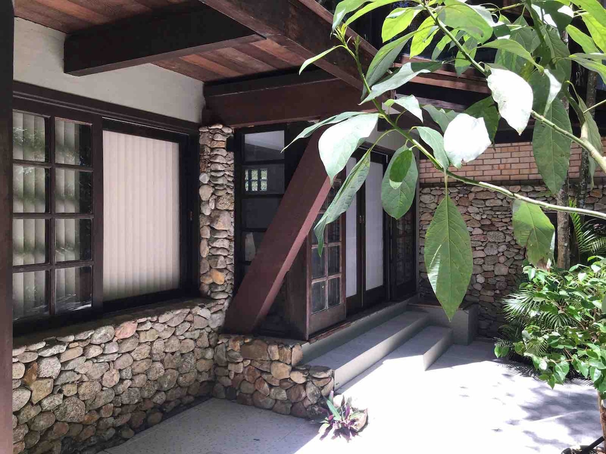 A charming exterior showcases a stone facade with wooden beams and large windows. Lush greenery surrounds the entryway, providing a serene environment. The pathway leads to a set of glass doors, inviting natural light into the space.