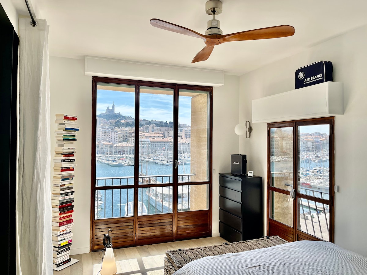 A bright bedroom showcases large windows with stunning views of the Vieux-Port and Notre Dame de la Garde. A tall bookshelf filled with books occupies one corner, while a dresser and a nightstand complement the space. Light wood flooring enhances the airy atmosphere.