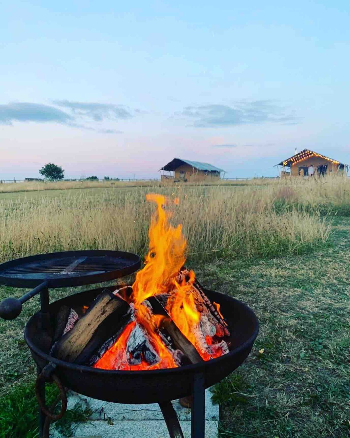 A firepit with bright flames provides warmth against a backdrop of open fields. In the distance, a safari tent is nestled under a soft sky that transitions from blue to pink as the sun sets.