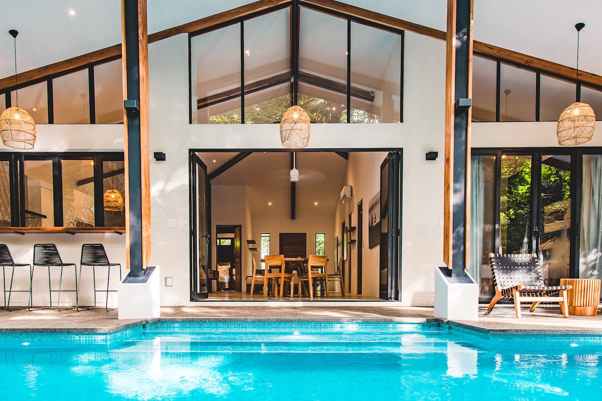 The image captures a serene view of a saltwater pool with clear turquoise water, framed by a modern home featuring large glass doors. The open-concept interior contains a dining area and kitchen, visible through the doors, while pendant lights hang above stylish bar seating.