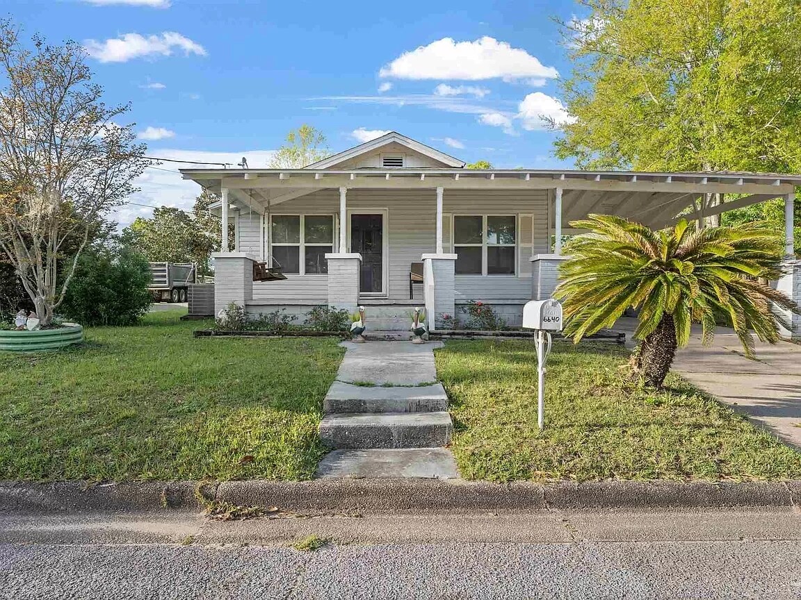 The front exterior of the home features a welcoming porch with a swing and several potted plants. A grassy yard surrounds the house, complemented by a palm tree on the right. The path leads to a white mailbox at the edge of the sidewalk.
