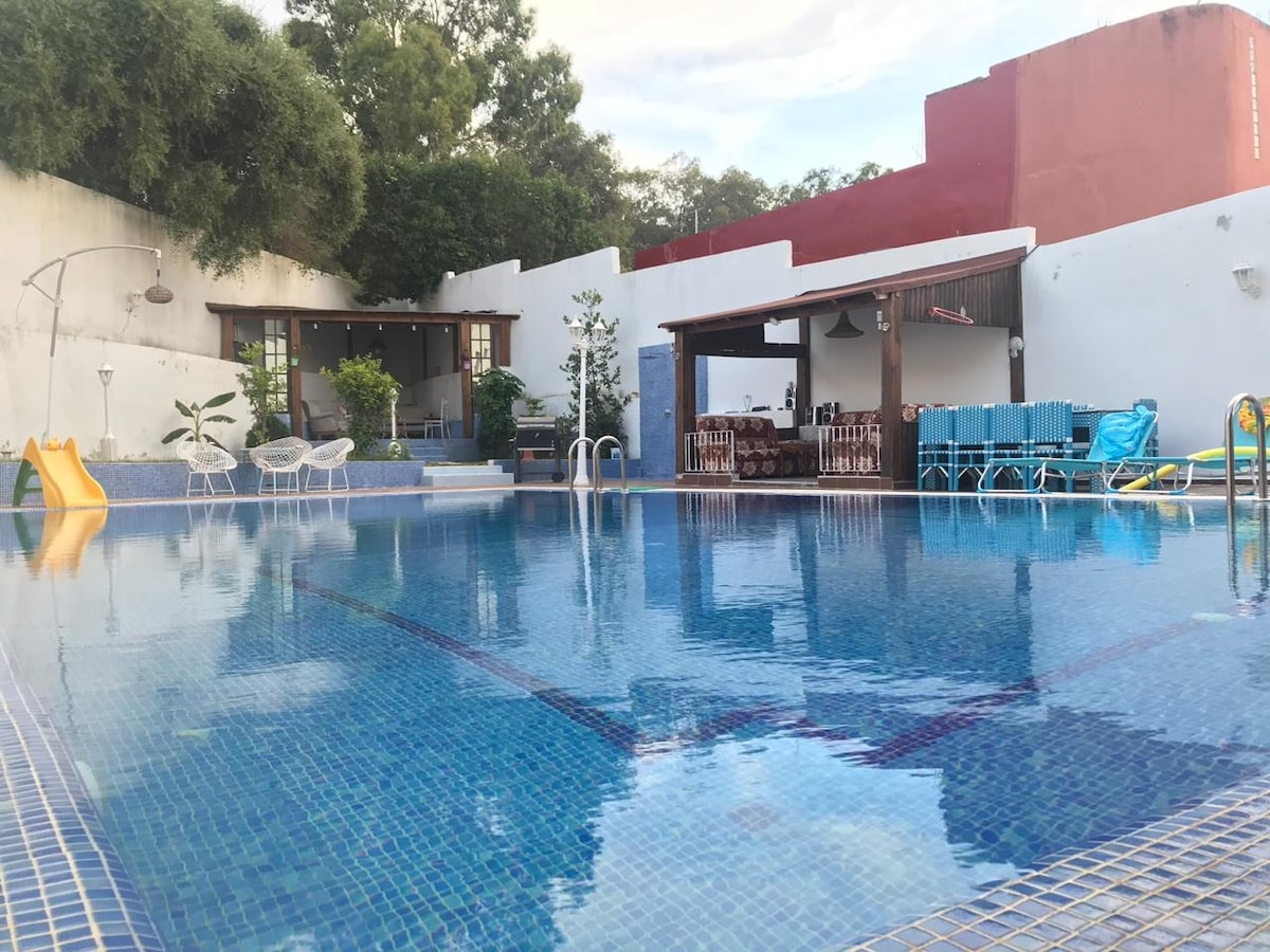 A private pool is framed by a spacious terrace, featuring lounge chairs and tables for relaxation. A bright yellow slide adds an element of fun, while greenery surrounds the area, creating a serene outdoor environment. The background showcases a covered seating space and open doors leading inside.